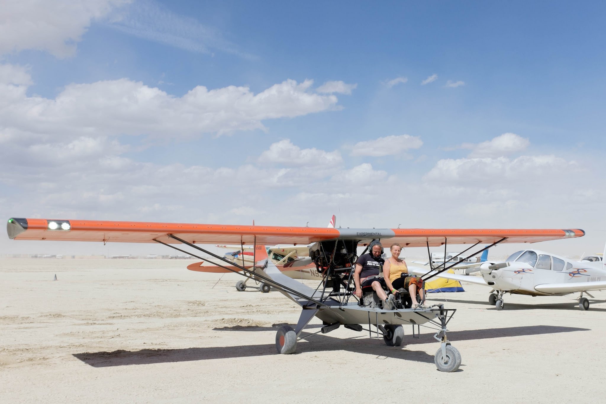 N149X, an orange-winged open-cockpit Breezy, parked on a dry lakebed with pilot and passenger in the seats